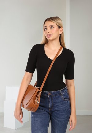 Schwarzes, geripptes, figurbetontes Oberteil mit V-Ausschnitt, kombiniert mit blauen, hochgeschnittenen Jeans. Braune Leder-Umhängetasche mit Klappe und verstellbarem Riemen.