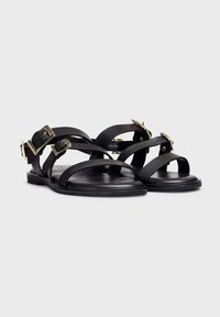 Sandalias de cuero negro con múltiples correas estrechas, hebillas en tono dorado y suela plana. Diseño simple con acentos mínimos y una textura suave.