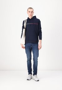 Mørkeblå hettegenser med en rød og hvit stripe over brystet, kombinert med lyseblå jeans og en beige totebag. Hvite joggesko.