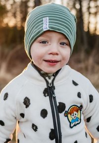 Valkoinen fleece-takki, jossa on mustia pilkkuja, vetoketjukiinnitys; vihreä raidallinen pipo, jossa on etiketti; takin päällä hahmopatchi.