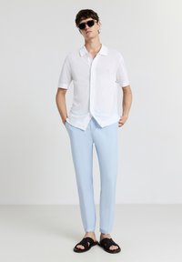 Weißes kurzärmeliges Button-Up-Hemd mit strukturiertem Muster, hellblaue maßgeschneiderte Hose und schwarze Sandalen mit offenem Zeh. Schwarze Sonnenbrille getragen.