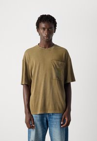 Mand iført olivengrøn oversized T-shirt med frontlomme og lyseblå jeans står foran en ensfarvet hvid baggrund.