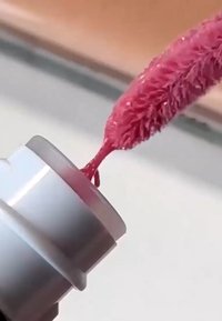 Gros plan d'un applicateur de gloss à lèvres rose prélevant une couleur à lèvres brillante d'un tube argenté sur un fond neutre.
