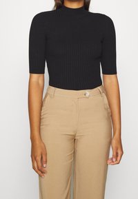 Haut noir à col roulé côtelé, à manches courtes, associé à un pantalon beige taille haute avec un détail de bouton unique et une texture lisse.