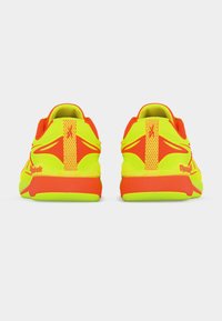 Φωτεινά κίτρινα και πορτοκαλί αθλητικά παπούτσια, εμφανιζόμενα από πίσω, με υφασμένα πέλματα και ταμπελάκια με το λογότυπο στη φτέρνα, σε λευκό φόντο.