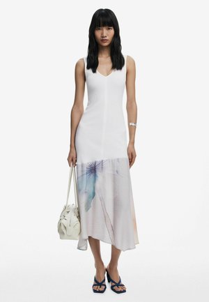 Weißes ärmelloses Kleid mit gerippter Textur und einem fließenden, gemusterten Saum in sanften Farben und floralem Design. Das Modell hält eine sandfarbene Handtasche.