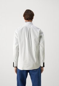 Weißes Hemd mit Knopfleiste, strukturiertem Kragen und kontrastierenden schwarzen Manschetten, kombiniert mit blauen Jeans. Der Stoff wirkt glatt und knusprig.