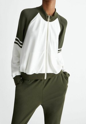 Person iført en hvid og olivengrøn lynlåstrøje med striber på ærmerne, matchende olivengrønne bukser, hænderne i lommerne.