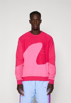 Pink sweatshirt with a textured design featuring overlapping shades of pink. Crew neck and long sleeves, paired with light blue pants.