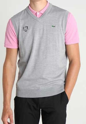 Homme portant un gilet en laine gris sans manches sur un polo rose à manches courtes avec un pantalon noir, debout avec une main dans la poche.