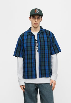 Jeune homme portant une casquette verte Salomon, une chemise à manches courtes à carreaux bleus et noirs par-dessus un t-shirt blanc à manches longues avec un motif de crâne, et un jean bleu.