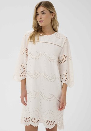 Mujer con un vestido blanco hasta la rodilla con bordes festoneados y bordado de ojales, de pie contra un fondo claro y liso.