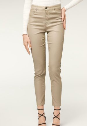Pantalon beige taille fine porté avec un haut blanc à manches longues et des sandales noires à talons hauts et à lanières, vus de la taille aux chevilles.
