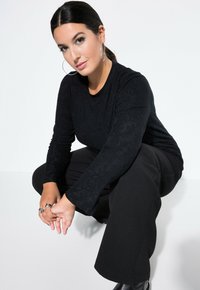 Schwarzes Langarmoberteil aus strukturiertem Stoff, kombiniert mit schwarzen weiten Hosen. Das Modell hockt und zeigt silberne Creolen und Ringe.