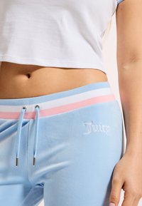 Gaiši zili velūra sporta bikses ar rozā un baltu svītrotu jostu, kurām sānos ir baltā izšūtā "Juicy" logo.