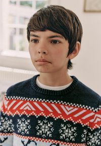 Sweter dzianinowy w kolorze granatowym z czerwonymi, białymi i jasnoniebieskimi geometrycznymi wzorami oraz motywami płatków śniegu; klasyczny dekolt typu crew neck.