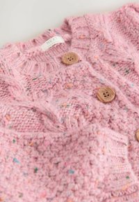 Różowy sweter z dzianiny z wzorem w warkocze, dwoma drewnianymi guzikami i kolorowymi drobinkami w całej tkaninie. Materiał jest miękki i fakturowany.