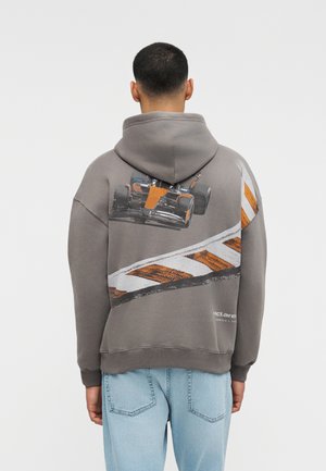 Άνδρας που φορά γκρι φούτερ με κουκούλα με γραφικό αγωνιστικού αυτοκινήτου σε πορτοκαλί και λευκό χρώμα και ανοιχτό μπλε τζιν, κοιτάζοντας προς τα πίσω σε απλό φόντο.