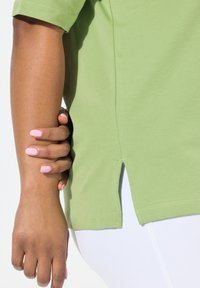 Grön bomullströja med korta ärmar, med sidsprund i fållen för ökad rörelsefrihet. Texturen verkar slät och mjuk.