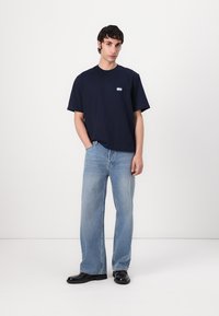Navy blue cotton t-shirt with short sleeves and a small logo patch. Paired with light blue wide-leg jeans and black shoes. Smooth texture.