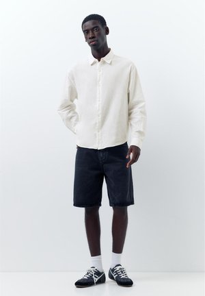 Junger Mann steht mit der rechten Hand in der Tasche, trägt ein weißes Hemd mit Knöpfen, schwarze knielange Shorts, weiße Socken und schwarze Turnschuhe.