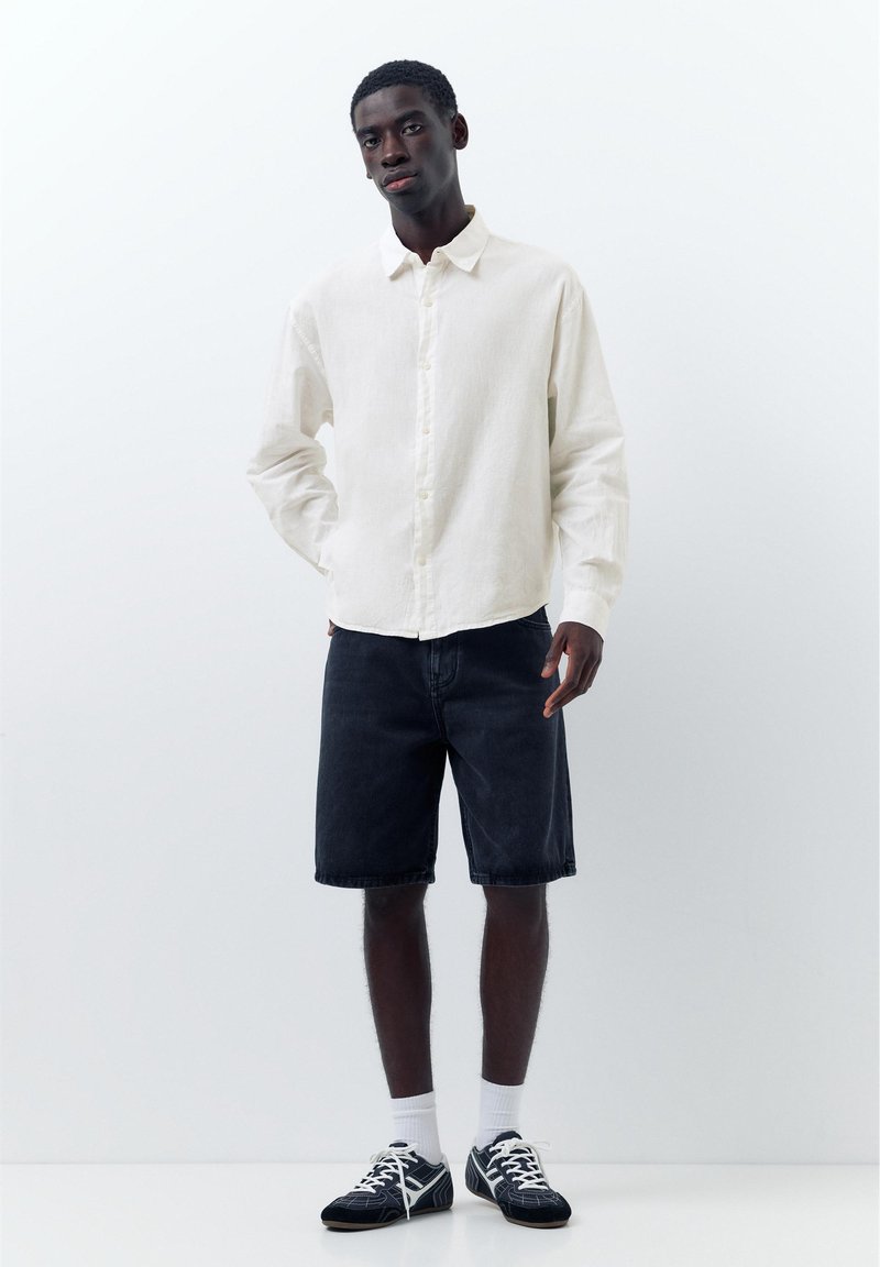 Jeune homme debout, main droite dans la poche, portant une chemise blanche boutonnée, un short noir jusqu'aux genoux, des chaussettes blanches et des baskets noires.