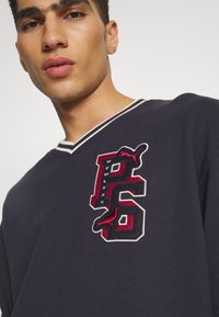 Jūras zilas v-veida kreklis ar baltām strīpām apkaklē, ar lielu izšūtu logo sarkanā un melnā krāsā priekšā.