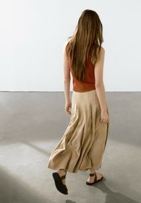 Gonna a pieghe color tan, top aderente ruggine e sandali neri. Lunghi capelli castani che fluiscono, girata di spalle su un pavimento liscio di un grigio chiaro.