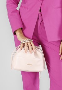 Borsa in pelle rosa chiaro di forma rotonda, con chiusura a coulisse e dettagli in tonalità oro. Posizionata in una mano con un bracciale a catena.