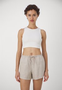 Bijela rebrasta crop majica u kombinaciji sa svijetlosmeštim lanenim kratkim hlačama s vezicom u struku. Model stoji na jednostavnoj pozadini.