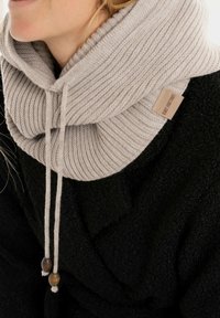 Lichtgrijze geribbelde gebreide col met koordjes en houten kralenaccenten, gedragen over een gestructureerd zwart kledingstuk. Etiket leest "Mint & Factory."