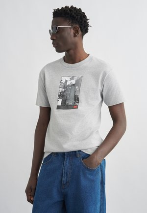 Jeune homme portant un T-shirt gris avec un motif d'immeuble urbain en noir et blanc et un jean bleu, debout avec une main dans la poche et des lunettes de soleil.