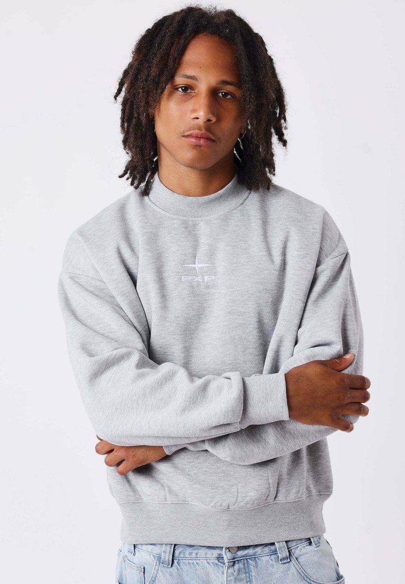 Jeune homme avec des dreadlocks portant un sweat-shirt gris clair avec un logo brodé et un jean bleu clair, debout les bras croisés sur fond blanc.