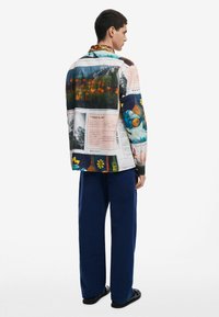 Camicia multicolore con stampe grafiche di natura e arte, colletto aperto e maniche lunghe. Abbinata a pantaloni blu larghi.