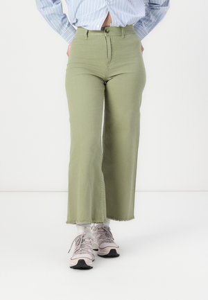 Pantaloni flare in denim verde oliva a vita alta con orlo grezzo. Indossati con sneakers rosa chiaro e calzini bianchi alla caviglia.