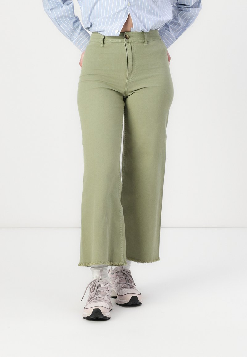 Pantaloni flare in denim verde oliva a vita alta con orlo grezzo. Indossati con sneakers rosa chiaro e calzini bianchi alla caviglia.