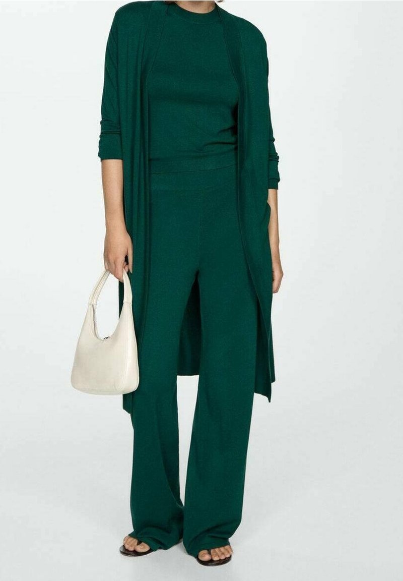 Tmavě zelený pletený outfit se štíhlým topem a širokými kalhotami, doplněný krémovou kabelkou na rameno a černými sandály.