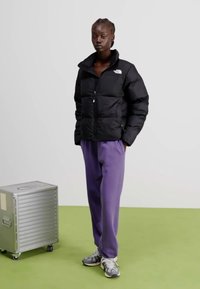 Giacca a vento nera con zip frontale e colletto arrotondato, abbinata a pantaloni da tuta viola. Un contenitore argento è vicino su un pavimento verde.