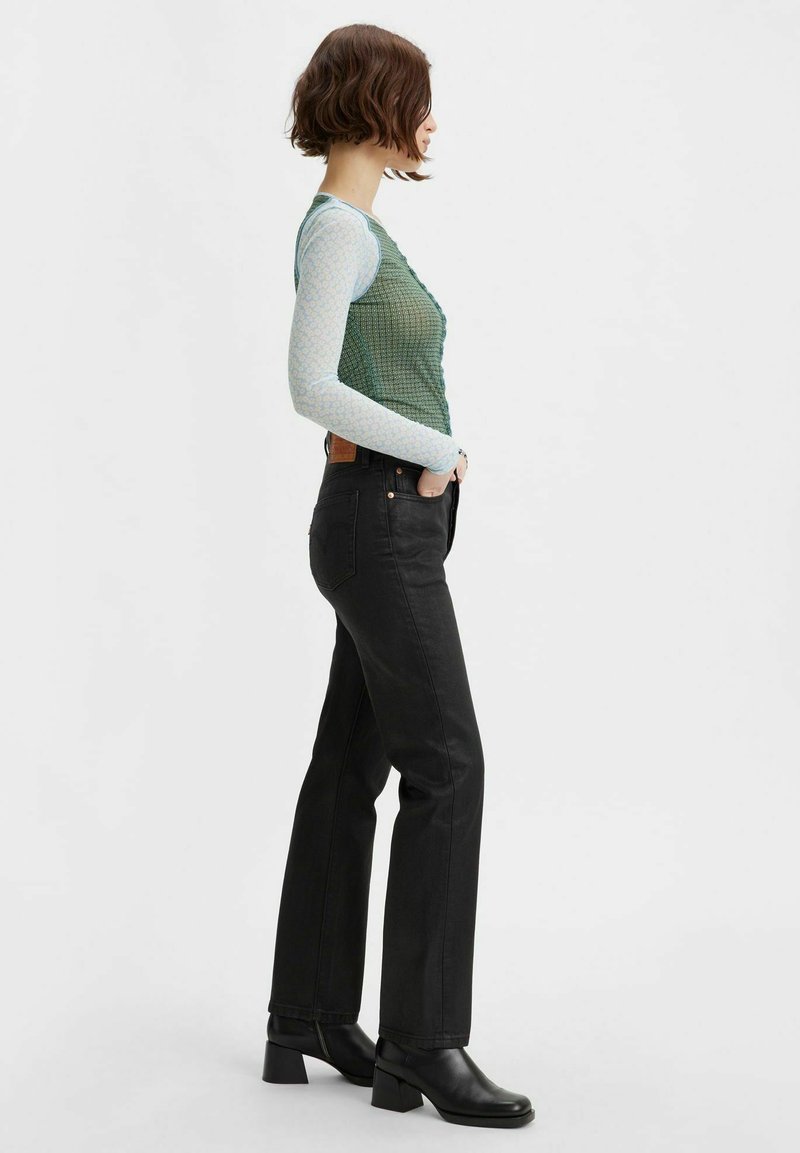 Gilet texturé vert superposé à un tee-shirt à manches longues bleu clair, associé à un jeans noir droit taille haute et des bottes noires à talons carrés.