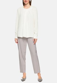 Weiße Bluse mit langen Ärmeln und plissiertem Vorderteil, kombiniert mit hellgrauen, maßgeschneiderten Hosen und grauen Ballerinas mit dekorativer Schnalle.