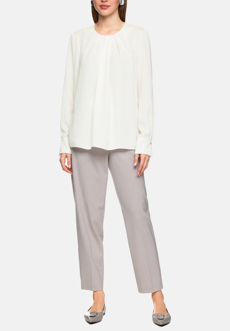 Weiße Bluse mit langen Ärmeln und plissiertem Vorderteil, kombiniert mit hellgrauen, maßgeschneiderten Hosen und grauen Ballerinas mit dekorativer Schnalle.