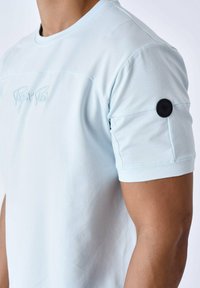 Lichtblauw T-shirt met korte mouwen van zachte stof, met een geborduurd logo en een zwarte cirkelaccent op de mouw.