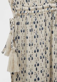 Tela beige clara con patrón de gotas en azul y negro; presenta borlas texturizadas en los lazos y un diseño de cintura fruncida.