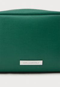 Πράσινη δερμάτινη τσάντα με λεία υφή και στρογγυλή μορφή. Διαθέτει μια ασημένια πλακέτα λογότυπου μπροστά και μινιμαλιστικό σχέδιο.