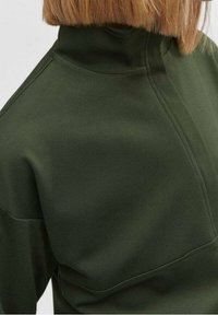 Jersey de cuello alto en verde oscuro con una textura suave y lisa; presenta una costura en el centro delantero y un diseño ajustado.