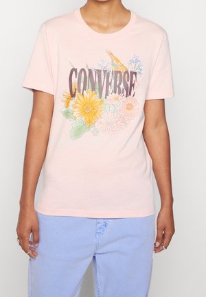 Personne portant un t-shirt rose pâle avec le texte "CONVERSE" et des motifs floraux jaunes et pastel, associé à un pantalon en jean bleu clair.