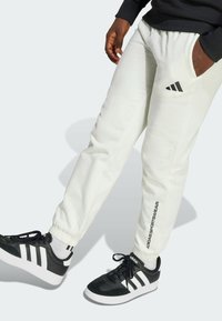 Bijele trenirke s elastičnim manžetama, s crnim Adidas logotipom na butu i bočnim natpisom. Nosene s crnim Adidas tenisicama.