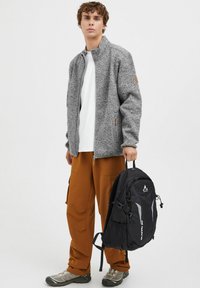 Jachetă gri din fleece cu guler înalt, pantaloni cargo bej și rucsac negru cu accente reflectorizante. Purtați cu pantofi sport gri.
