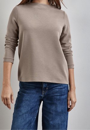 Vrouw draagt een beige geribde top met lange mouwen en geplooide mouwen, gecombineerd met donkerblauwe spijkerbroek tegen een effen witte muur.