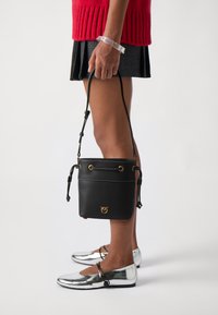 Μαύρη δερμάτινη τσάντα τύπου bucket με χρυσές λεπτομέρειες, κλείσιμο με κορδόνι και υφή. Συνδυασμένη με λαμπερά ασημί φλατ παπούτσια και πλεκτό πουλόβερ.